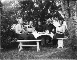 Fire damer, et barn og en mann sitter ved et bord i hagen. P