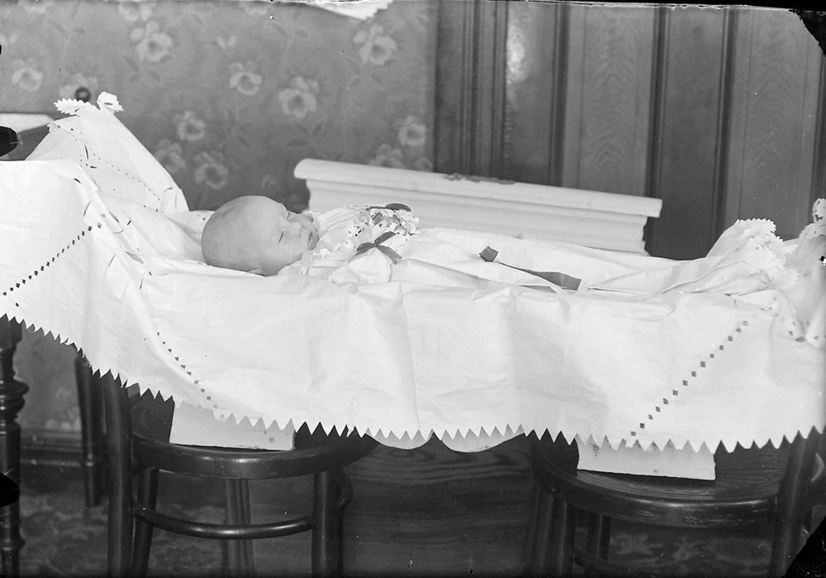 Begravelse. Et lite barn ligger i en åpen kiste. Aminda Marie Olsen, f. 1914, d. 1915. Datter av Oluf Sidenius Norman Amundsen/Olsen og Katinka Marie Bergljot Nilsen, Reine, Moskenes.