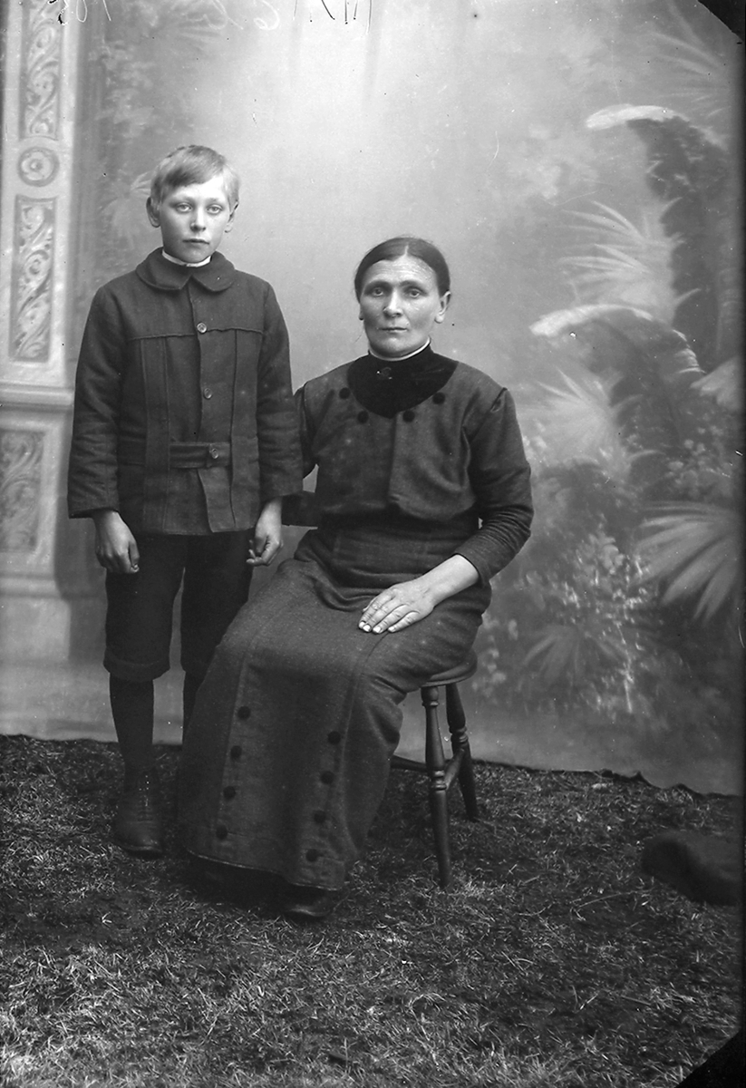 Portrett. En gutt og en kvinne. Kvinnen sitter på en stol, gutten står ved siden av. Kvinnen har kjole på seg, og gutten har bukse og jakke på seg.Bildet er tatt på Ramberg i Lofoten.