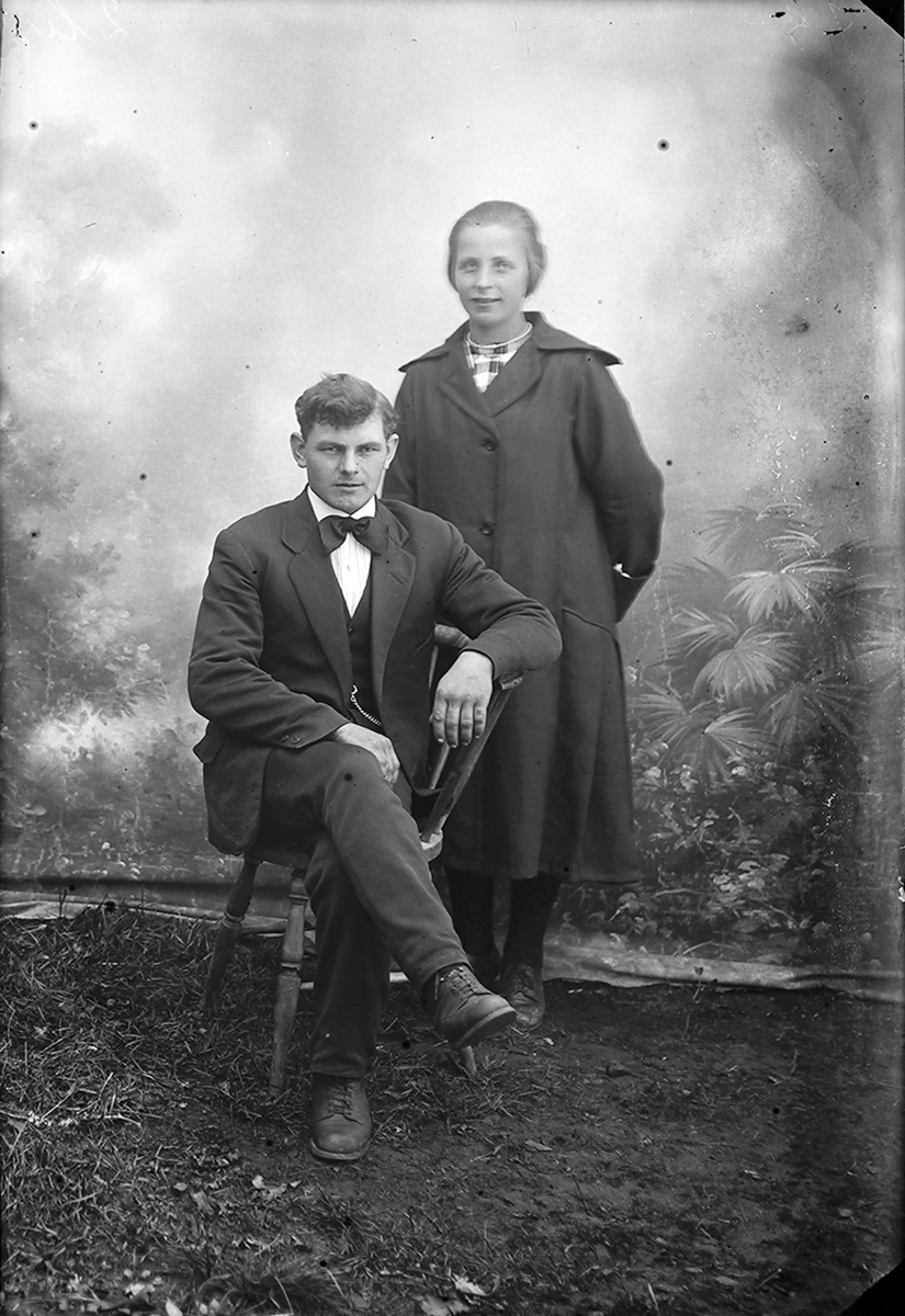 Portrett. En mann og en kvinn. Mannen sitter på en stol og kvinnen står bak. Mannen har på seg dress med sløyfe i halsen. Kvinnen har kåpe på seg. Sørfoldbilde.