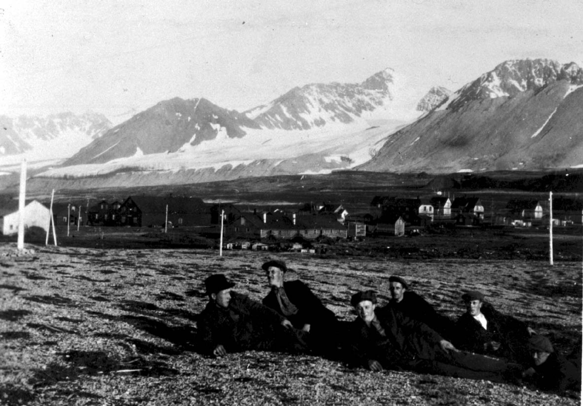 Gruppebilde, 5 personer, menn sitter/ligger på bakken. Bygninger og fjell i bakgrunnen.
