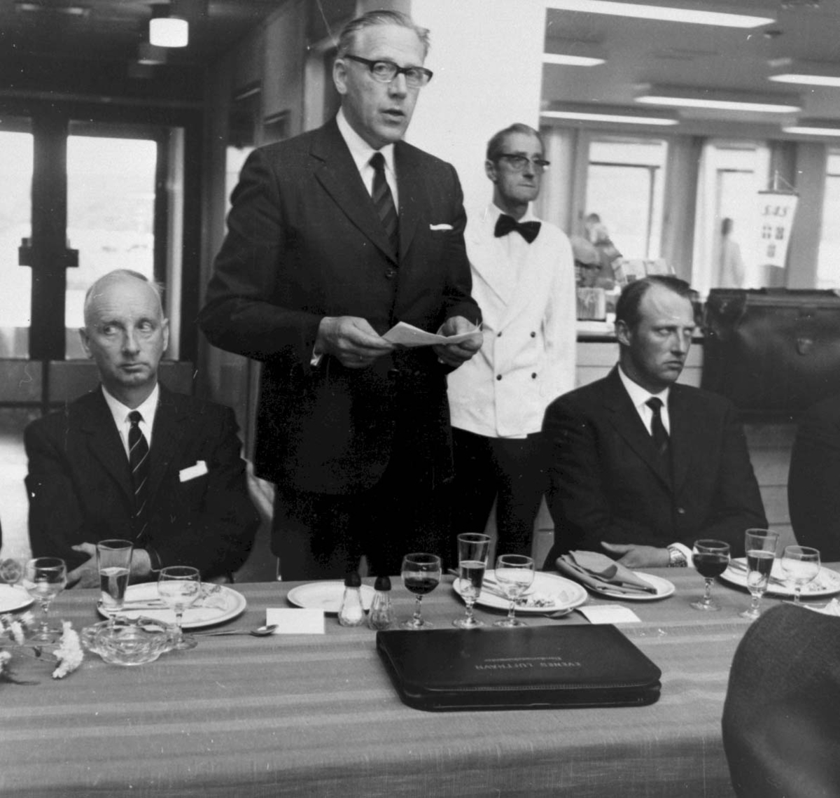 Lufthavn Gruppebilde, blant annet H.K.H Kronprins Harald sitter ved dekket bord. 1 person, mann holder tale.