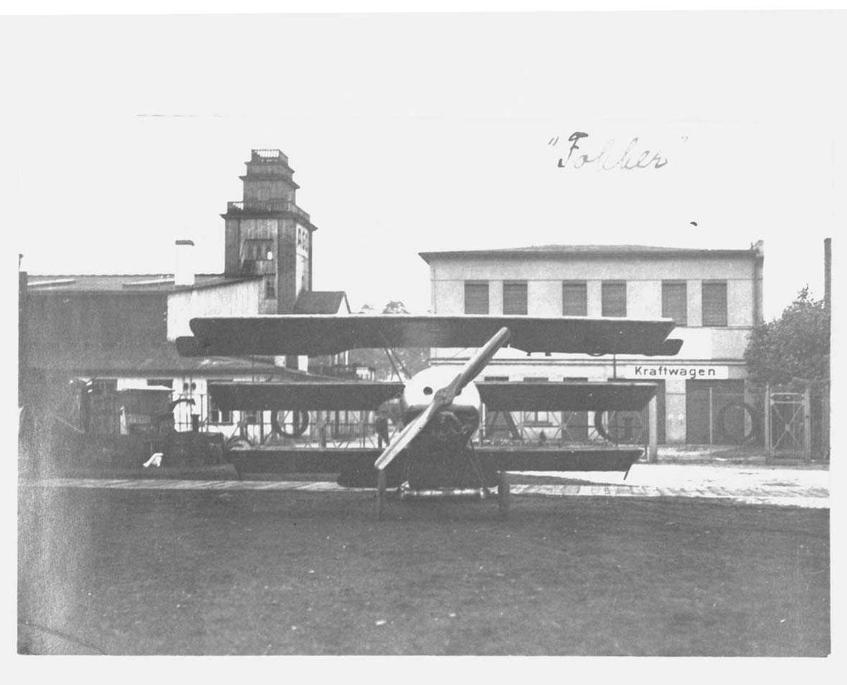 Ett fly på bakken.Fokker Dr 1, bygninger i bakgrunn, 1 person bak fly