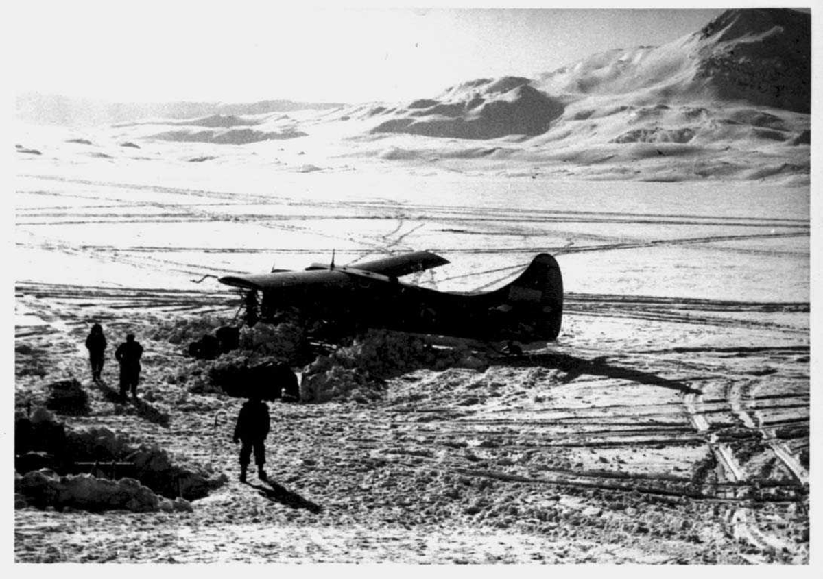 Snødekt landskap. Ett fly på bakken, Otter. Tre personer og noe utstyr ved flyet.
