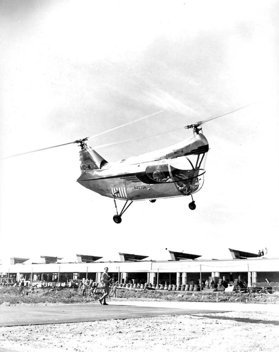 1 helikopter i luften. McCulloch MC-4. 1 person i cockpiten. Bygning med flere personer i bakgrunnen.