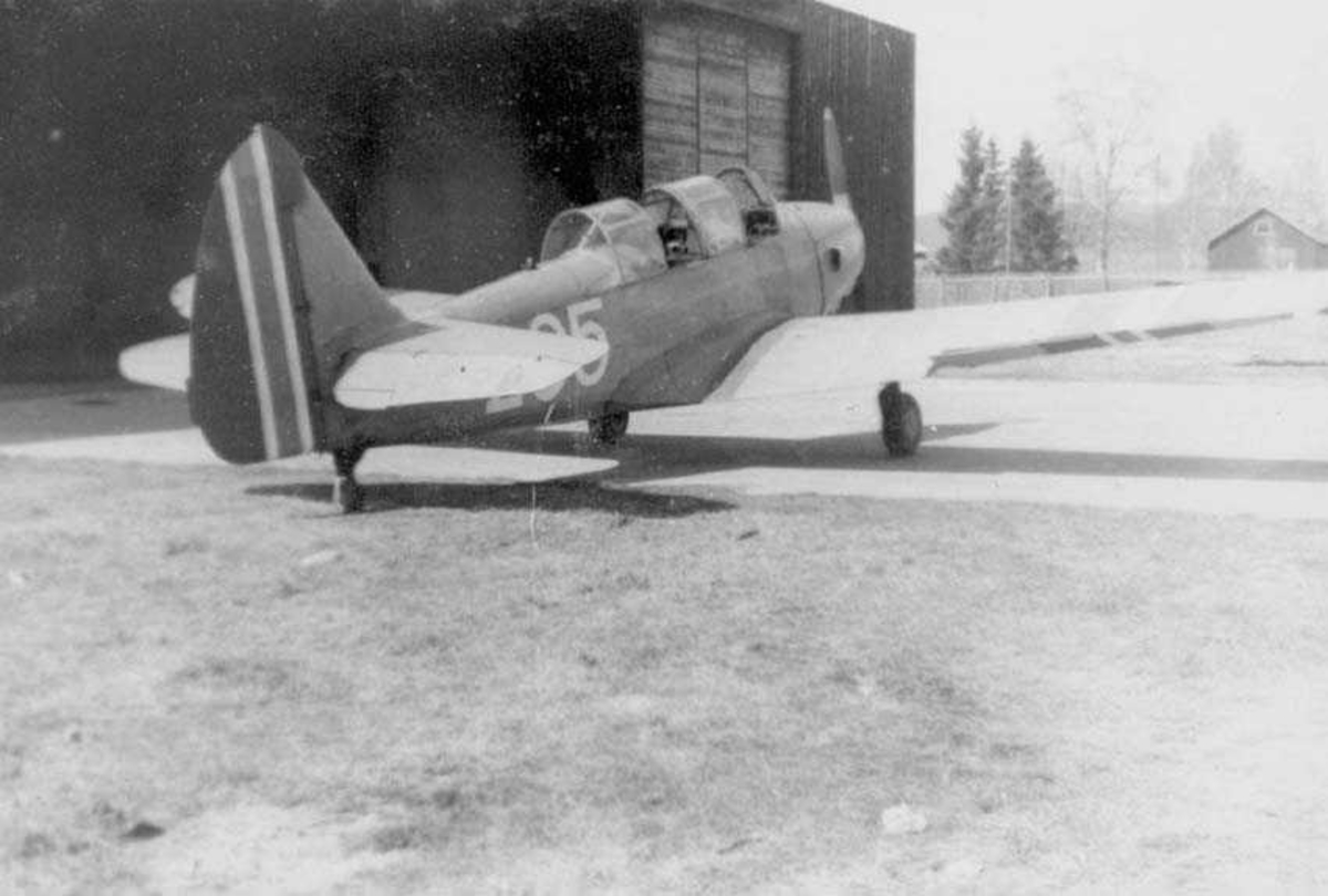 Et fly på bakken. Fairchild Cornell. Sett bakfra. Står utenfor en hangar. Bygning i bakgrunnen.