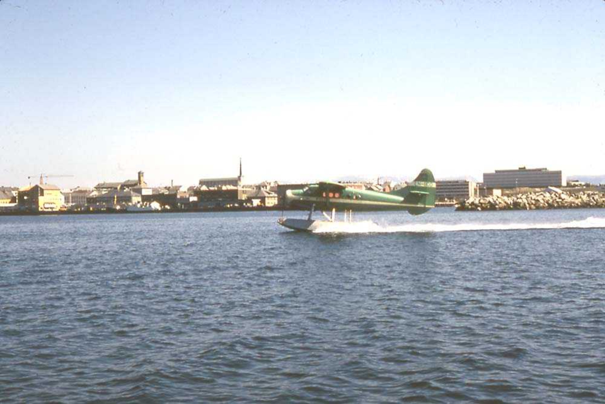 Ett fly i fart på vannet, DHC-3 Otter LN-BDD fra Widerøe. Bygninger/by (Bodø) i bakgrunnen.
