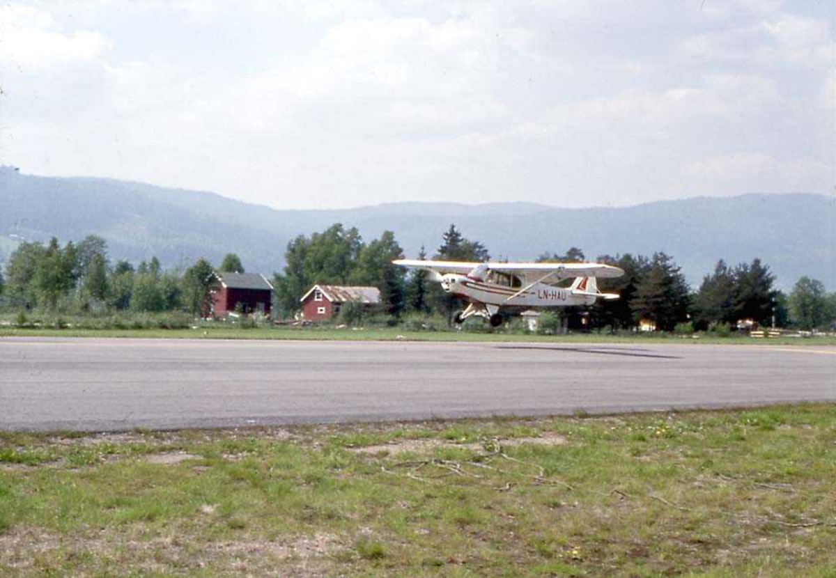 Ett fly like over bakken som tar av/lander, Piper PA-18-150 Super Cub, LN-HAU. Rullebane under flyet. Bygninger og fjell i bakgrunnen.