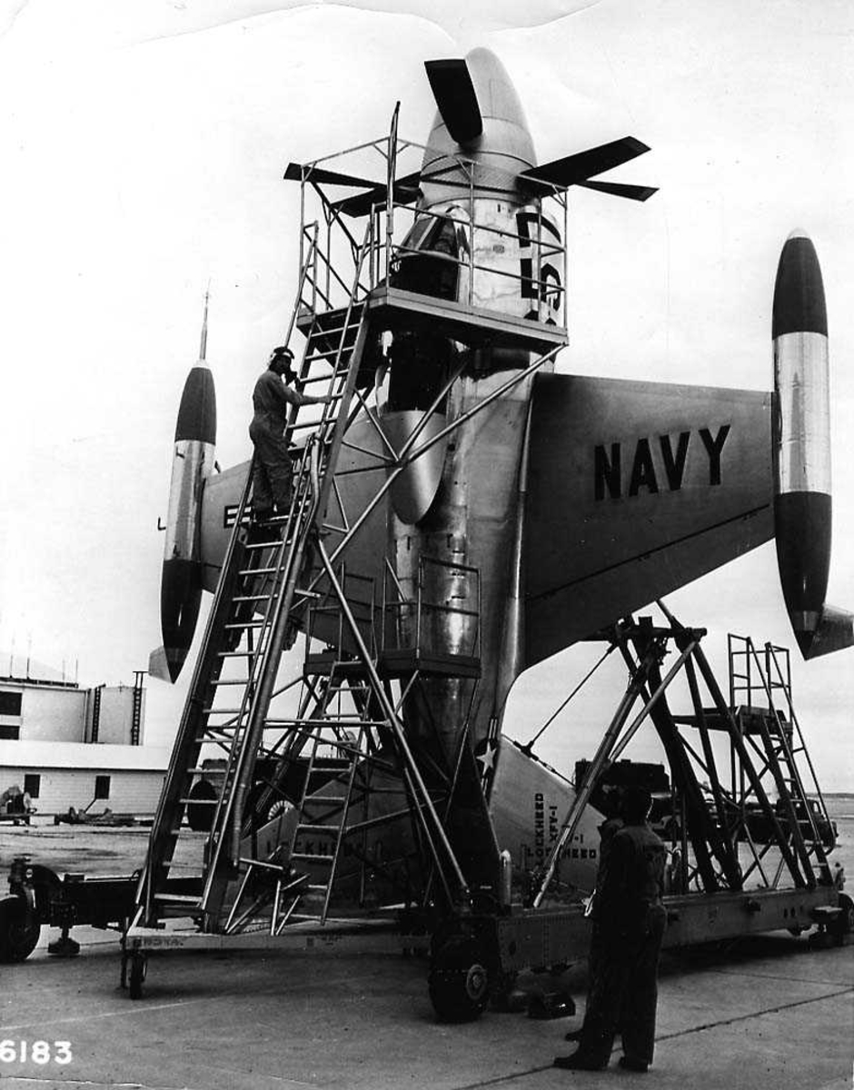 Ett fly på bakken som står vertikalt, Lockheed XFV-1. Et stativ står inntill flyet med en pilot som klatret opp en stige til cockpiten. To personer på bakken.