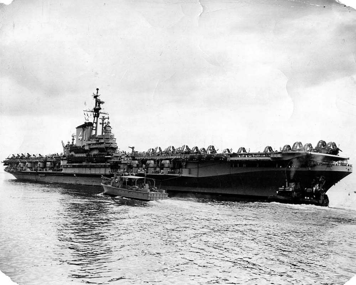 Ett hangarskip, USS Midway. Flere fly med oppslåtte vinger på dekket. En liten båt i sakte fart foran hangarskipet.