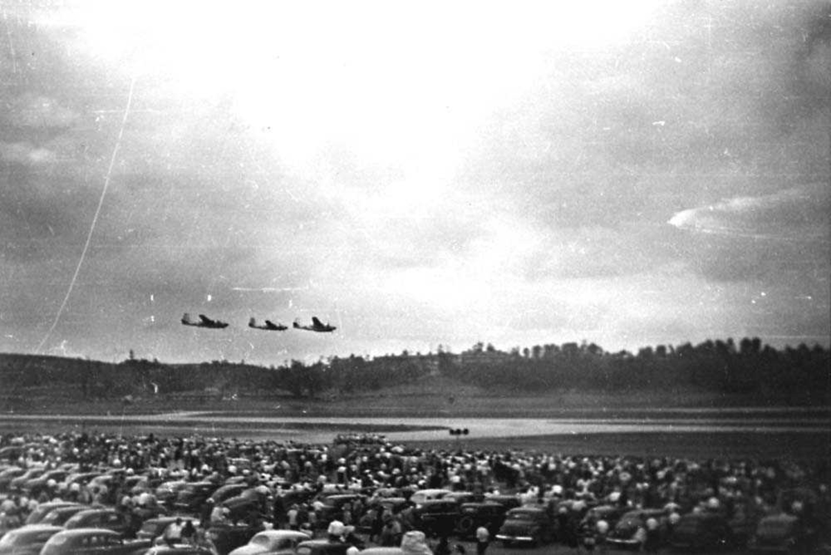 Tre fly i luften som flyr i formasjon foran en folkemengde som står på bakken. Mange biler blandt folkemengden.