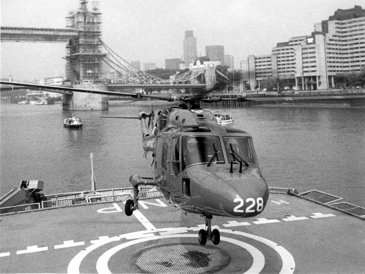 Ett helikopter i luften, like over skipsdekk/helipad. Westland Lynx Mk.86 tilhørende 337 skv. merket 228. Skipet ligger på elven Thames med Tower Bridge under opppussing bak t.v i bildet.