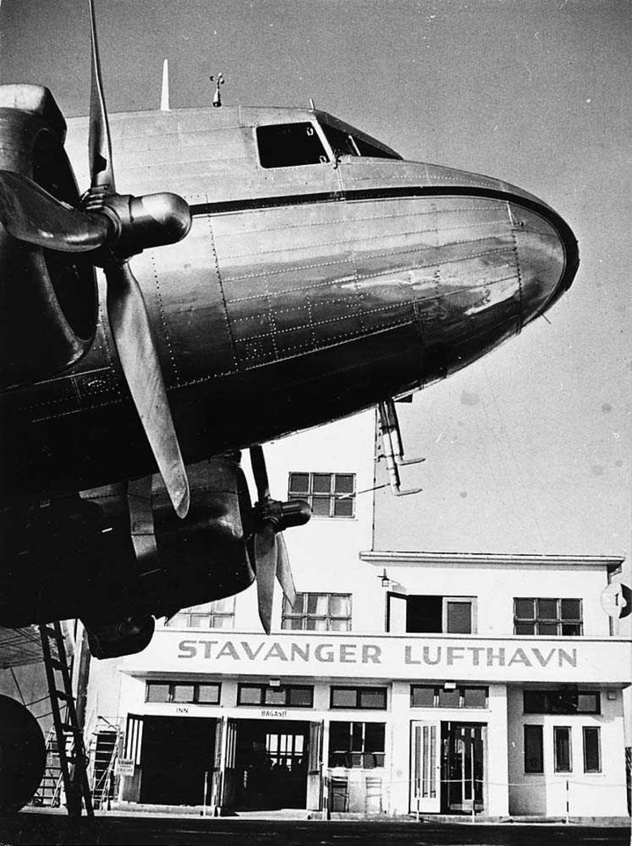 Lufthavn. Forparten til ett fly på bakken, Douglas DC-3/C-47 Dakota. Bygning i bakgrunnen.