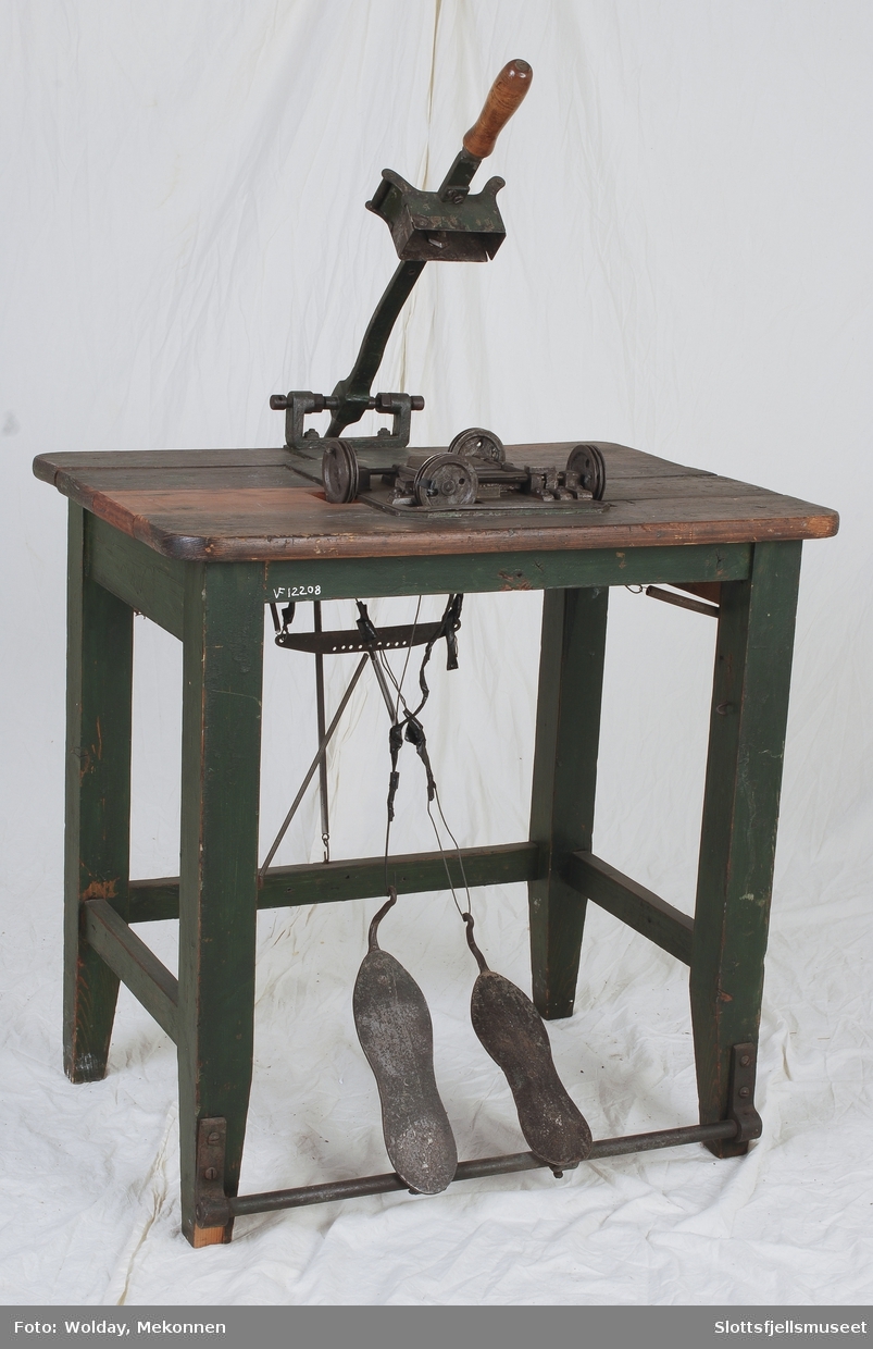 Trebord med påmontert mekanisme bestående av metallplater, hjul og stenger.  2 fotplater er forbundet med metalltråd opp til håndtak som kan dyttes ned mot bordet.