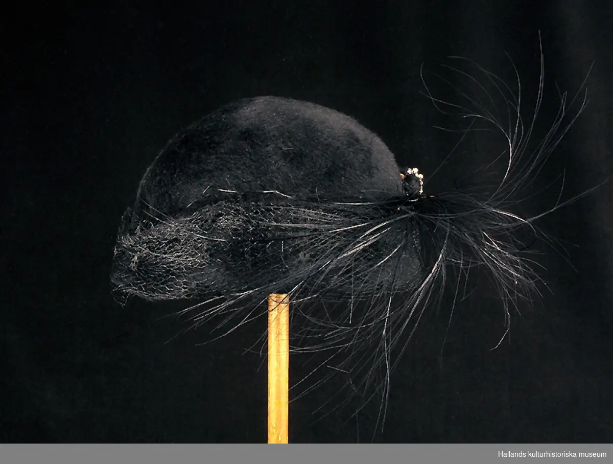 Damhatt av svart filt med plyschliknande utsida. Rund form, försedd med flor, en plym och ett hattsmycke i strass och pärlor. 
Varumärkt: "Asta Liljegren VARBERG TEL. 468"