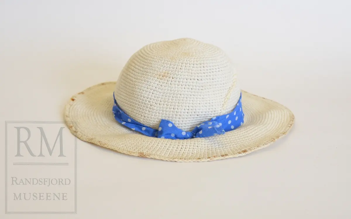 Sommerkjole m/hatt. a) Bomullstoff, blått med hvite prikker. Sammensydd i livet. Overdelen har Lukning foran med 5 hvite knapper og knapphull, puffermer. Krage med heppekant. 6 Bredder i utskrådd skjørt. Lukning til venstre for midt foran. 
b) Belte med knapp og knapphull. 
c) Hatt, Heklet av hvitt bomullsgarn. Ståltråd ytterst i Bremmen (rusten) Figurkant på pullen. Pyntebånd av kjoletøyet.