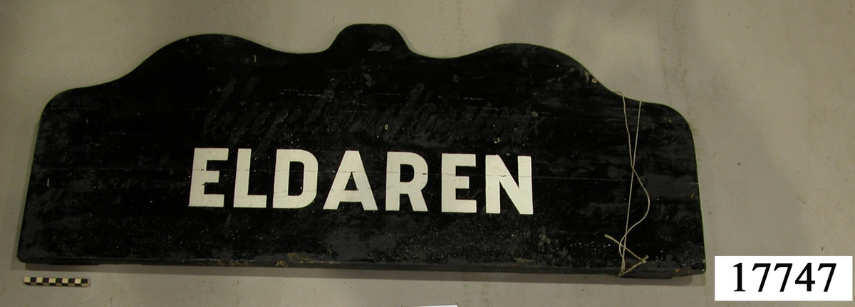 Rektangulär skylt, ovansidan sågad i tre bågar, svartmålad. Vit schablonmålad text: " ELDAREN ", övermålad text under svart färg: " Uppbördsrum ".