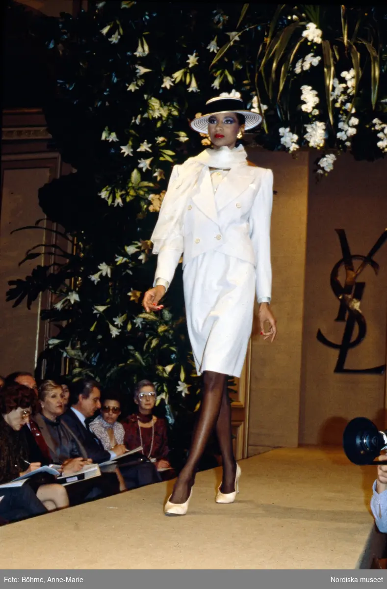 Modevisning. Modell i vit klänning, kavaj, sjal och hatt med hattband. Från Yves Saint Laurent.