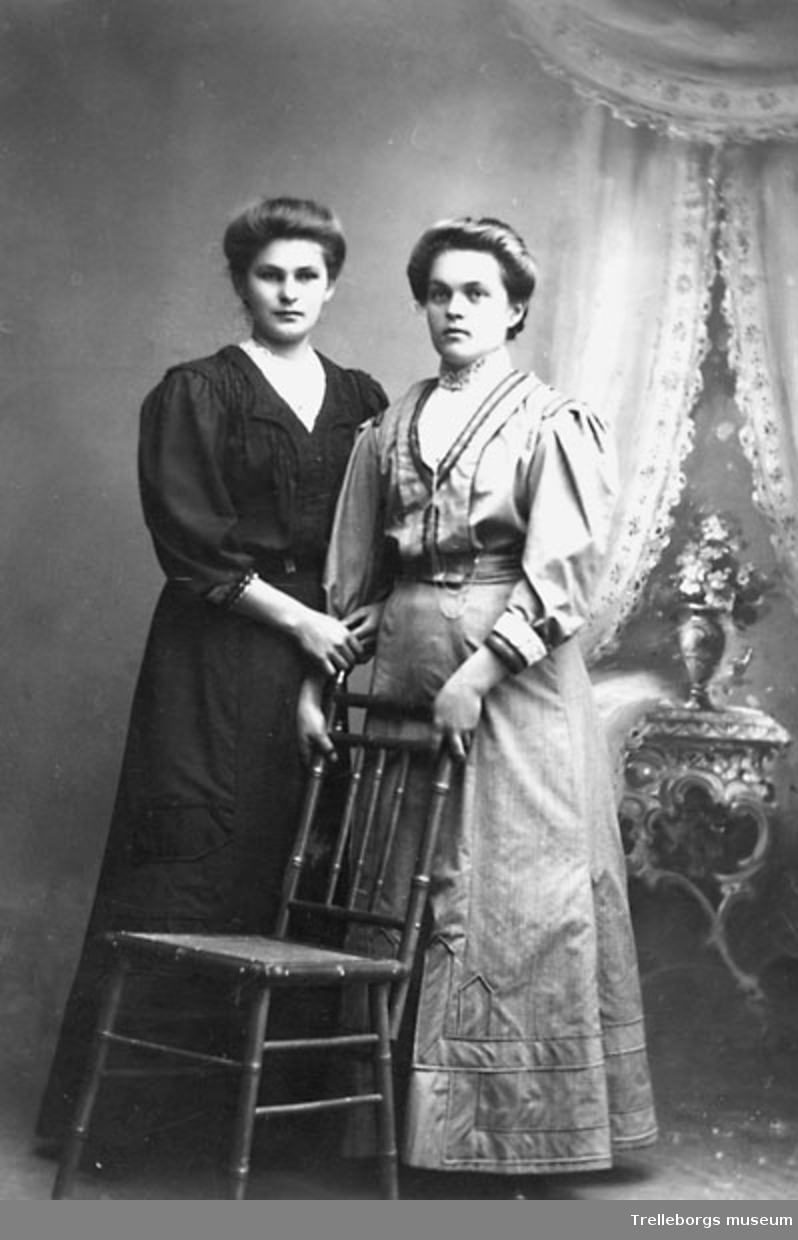 Två kvinnor, en i svart klänning med inslag av vitt och en i ljus klänning och med urkedja om halsen.