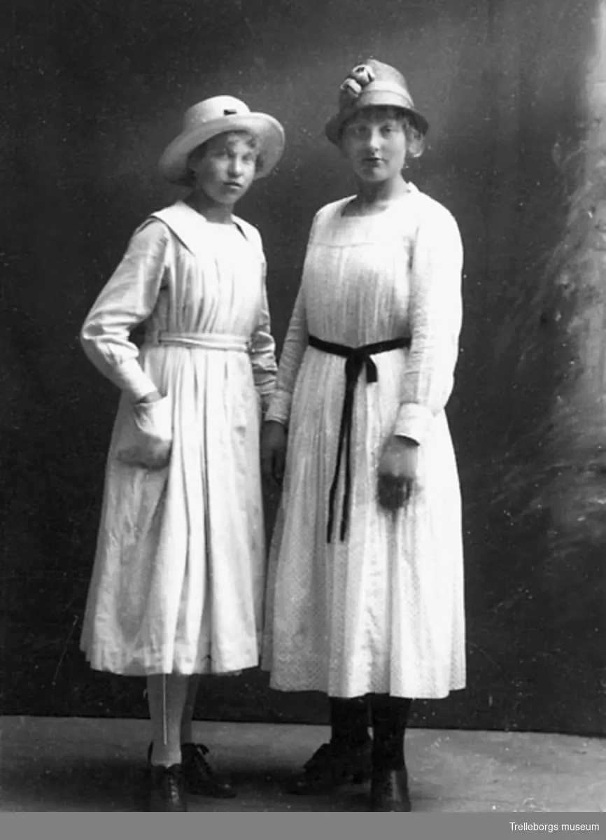 Två unga kvinnor i ljusa klänningar och halmhatt.
