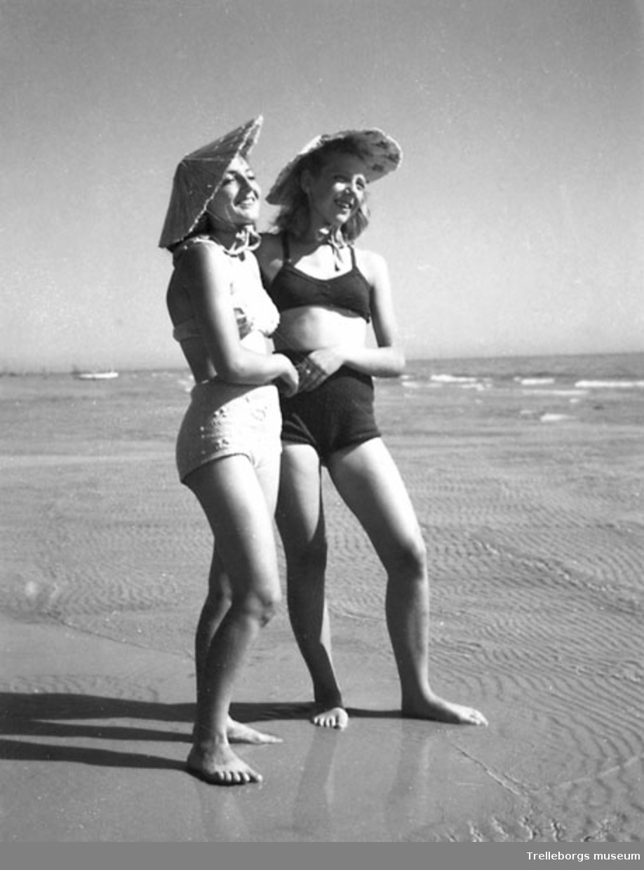 Två kvinnor i baddräkt och solhatt på stranden.