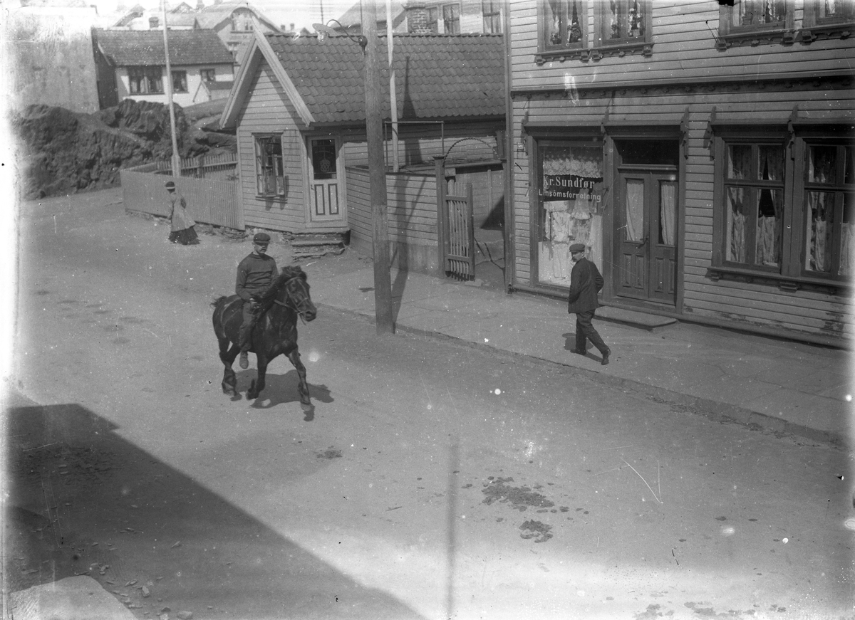Haraldsgaten ved Kolbeinshaugen. En mann ridende på en hest midt på gaten. En mann på fortauet på høyre siden. Trehus på høyre side og i bakgrunnen, i tillegg til et gjerde og en flaggstang og en annen person.
