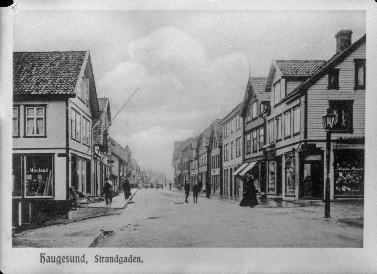 Strandgaten i Haugesund sett fra syd i 1905. "Morland" malt på vinduet på huset til venstre. Gaten midt på bildet. Gatelykt til høyre. Trehus på begge sider. Flere personer i gaten. Hest og kjerre i bakgrunnen.