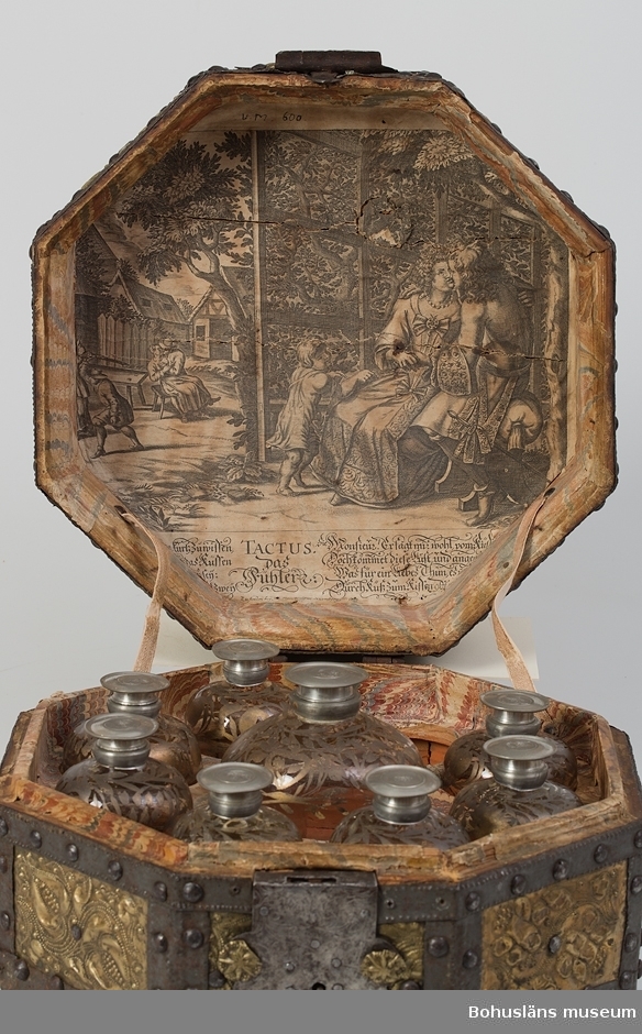 Ett åttkantigt skrin för nio glasflaskor. Skrinet är ett holländskt arbete från 1600-talet.
Skrinets utsida är beslagen med mässingsplåt, ca 0,2 mm tjock och hålls på plats med smidda band och prydnadsband av järn. Mellan banden framträder dekorativa rektangulära mässingsfält. Lockets två parallella band är klippta i ett löpande åttakantigt mönster passande skrinets form. Mässingsplåten är rikt dekorerad med blommor, hjärtan, cirklar och löpande linjer vilka formats med puns eller pressats fram.
Invändigt är skrinet rödbemålad i dess undre del med vad som troligen är tunn oljefärg, alternativt limfärg. Skrinets lock och den övre hälften av skrinets underdel är klistrat med papper av två slag. Locket pryds av ett kopparstick av amoröst slag som utgör en mindre del av en större bild. Texten "Tactus das fuhler", troligen motivets titel, är urklippt och placerad i anslutning till motivet. Ytterligare text beskriver kärlekens nöjen. Under titeln står "zu finden bey Joh. Peter Wolff in Nurnberg".
Övriga partier av ett marmorerat papper av holländskt slag. Marmoreringen är utförd med kam i rött, blått, vitt och gult. 
Lock och underrede hålls samman av två smidda gångjärn. I uppfällt läge har locket hållits på plats med två rosafärgade linneband.
Skrinets lås har låsbricka av järn dekorerat med ciselerade bladslingor.
Skrinet har kunnat bäras i ett handtag av smidesjärn, fästat ovanpå skrinets lock.
Inuti skrinet står sju mindre flaskor av blåst glas, (höjd 11 cm, diameter 7 cm) samt en större flaska (höjd 13 cm, diameter 10 cm). Flaskorna har svarvade och stämplade skruvlock av tenn. Flaskornas skuldror är dekorerade med bladguld lagt i olja och föreställer blad, blommor och torligen frukter.
Utförande är ganska grovt med obehandlade geringar, och sammanfogning med mindre spik.
Om Johann Peter Wolff finns uppgifter att han gjorde kopparstick på 1700-talets början.
Ur handskrivna katalogen 1957-1958:
8-kantigt skrin med 8 glasflaskor [en flaska saknas av de nio platser för flaskor som finns]. Skrinets L. 30,3 Br. 30,3 H. 16,5. Utvändigt klätt med järn- och kopparbleck [ska vara mässingsbleck]. Invändigt ställ för flaskor. Skrinet något skadat. Enstaka maskhål. En större flaska diam: 9,5. H.: 13. 
7 mindre flaskor. 5 st.: diam. 6,2. 2 st.: diam.: 6,0. Samtliga H:11. Med guldmönster och hattar av vitmetall. Hela.

Lappkatalog: 61
Ur handskriven katalog över Uddevalla - Musei Historiska - samlingar: Utländska föremål = De etnografiska föremålen upprättad  år 1916 av Knut Adrian Andersson, Intendent från 1 Oktober 1915:
No 22. Rese-Skrin, åttasidigt; utvändigt konstrikt beslaget med järn- och mässingsnaglar, inuti har det 8 glasflaskor med hattar av vit metall märkta L och däröver kunglig krona. Skänktes 1864 av Disponenten vid Kampenhofs fabrik i Uddevalla Robert Wilhelm Bley. (=G:23 i 1869 års tryckta katalog.)

Litteratur:
Framtidens Skrin, 300 års design, Hemslöjd Västra Götaland, s. 13 (Medicinskrin från 1600-talet; RMVG21609).
Wallin, Sigurd: Nordiska museets möbler från svenska herremanshem. Del I, 1500-1600-talen. Renässans och barock. Nordiska museet 1931 - 1935. Facsimile-upplaga 1979.