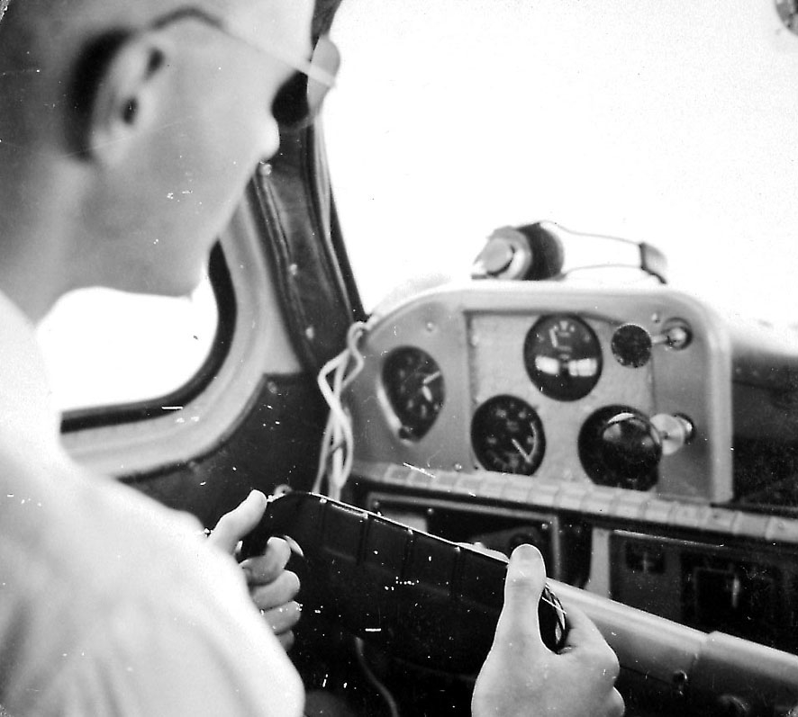 En person, flyger, sitter i cockpiten på et fly.  Noe av instrumentpanelet ses.
