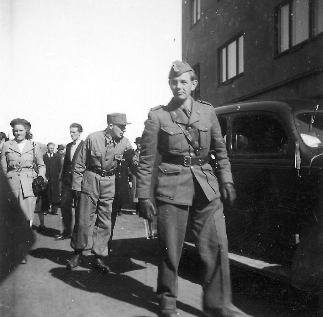 Frigjøringsdagene i Bodø etter krigen 1940 - 1945. To militer personer foran, flere sivile bak. Ett kjøretøy og noe av bygning til høyre.