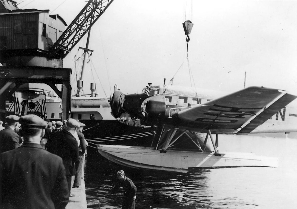 Ett fly, Junkers Ju 52/3mg2e, LN-DAH, "Falken". Henger i heisekran. Flere personer ved flyet. Litt av overbygg på en båt i bakgrunnen.