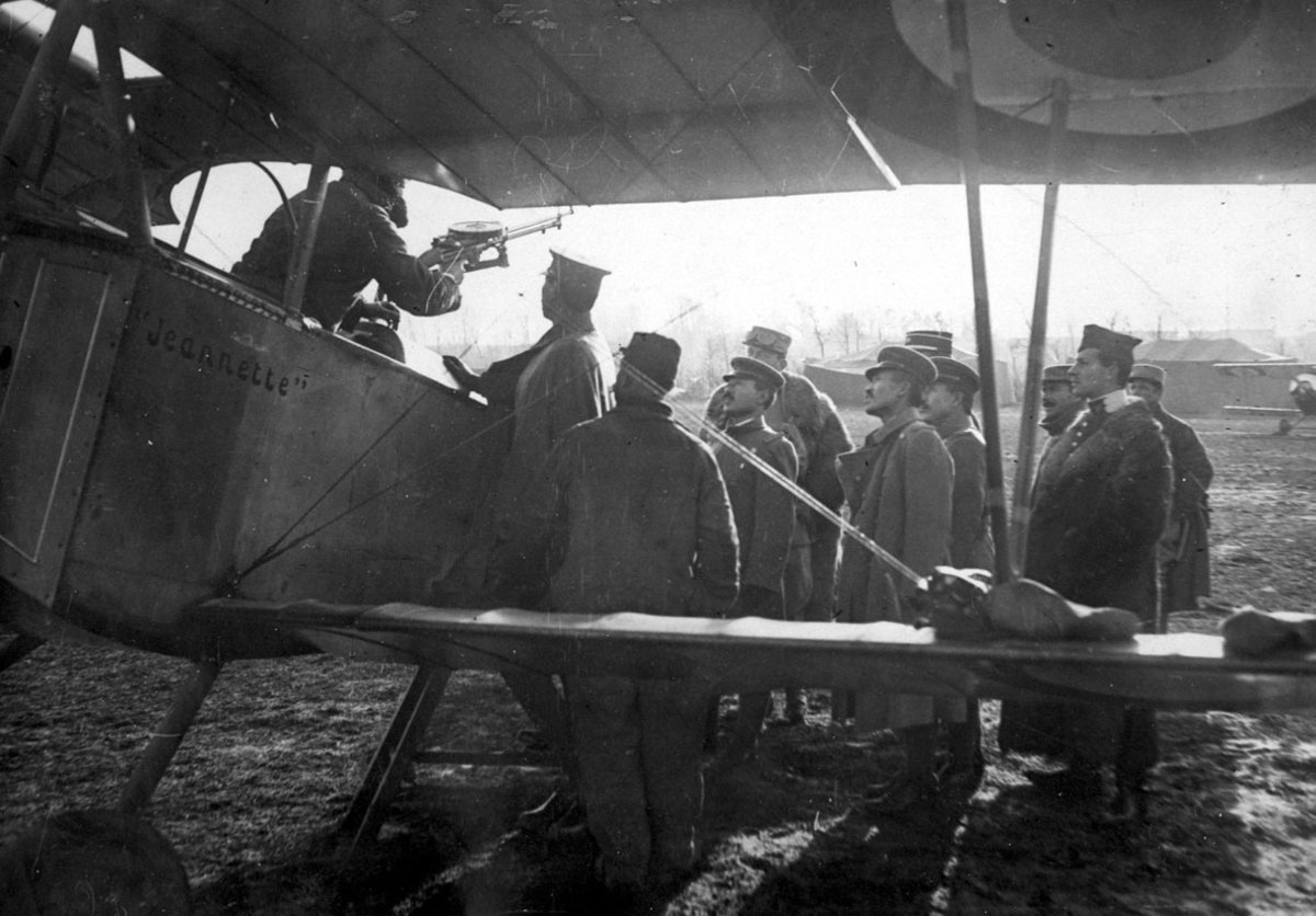 Fly, Nieuport 11C.1 "Jeanette", på bakken. Skrått forfra, midtpartiet. Flere personer ved flyet. 1 person i cockpit, "justerer" påmontert våpen, maskingevær.