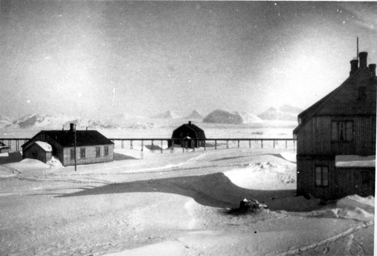 3 bygninger. Jernbanelinje på pæler i bakgrunnen. Snø på bakken