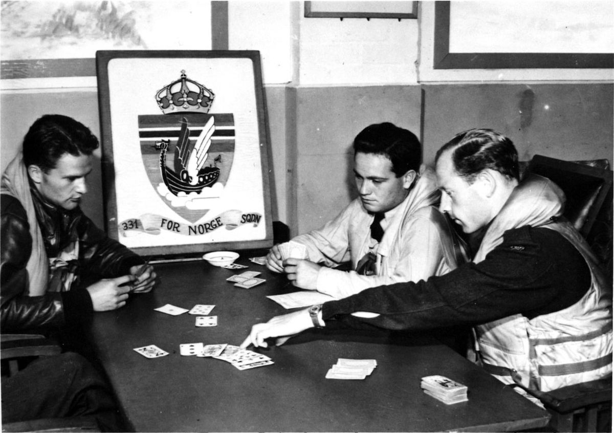 3 personer i militæruniform, 2 med pillotutstyr. Sitter og spiller kort rundt et bord. Logo på oppreist på bordet " Vikingeskip - 331 FOR NORGE SQDN"