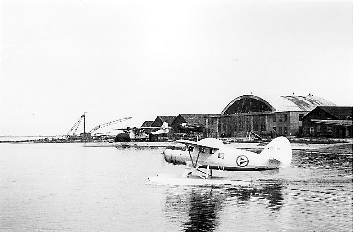 1 fly, Norsemann Mk.VI-UC-64-ND, i sakte fart på havoverflata, nært land. Flere bygninger bak. Lenger bak sees 2 andre fly, stå oppe på land, og en større heisekran.