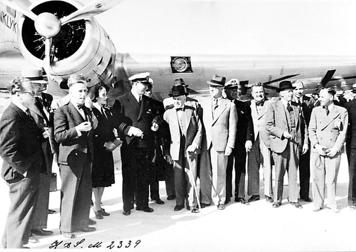Lufthavn, portrett, flere personer, noen i flyuniform, står oppstilt foran motoren på et fly DC-2 fra KLM. Tatt utendørs.