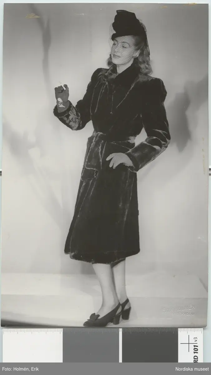 Nordiska Kompaniet. Kvinna i mörk päls med brett skärp och svart hatt med flor. Cigarett i ena handen. Text på baksidan: "Arkivbild d. 21/8 45"