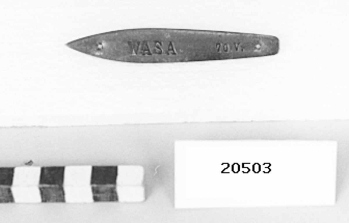 Skylt av mässingsplåt, i formen som fartyg med hål i för och aktern för upphängning. Instansad text på mitten: "WASA 70 V.".