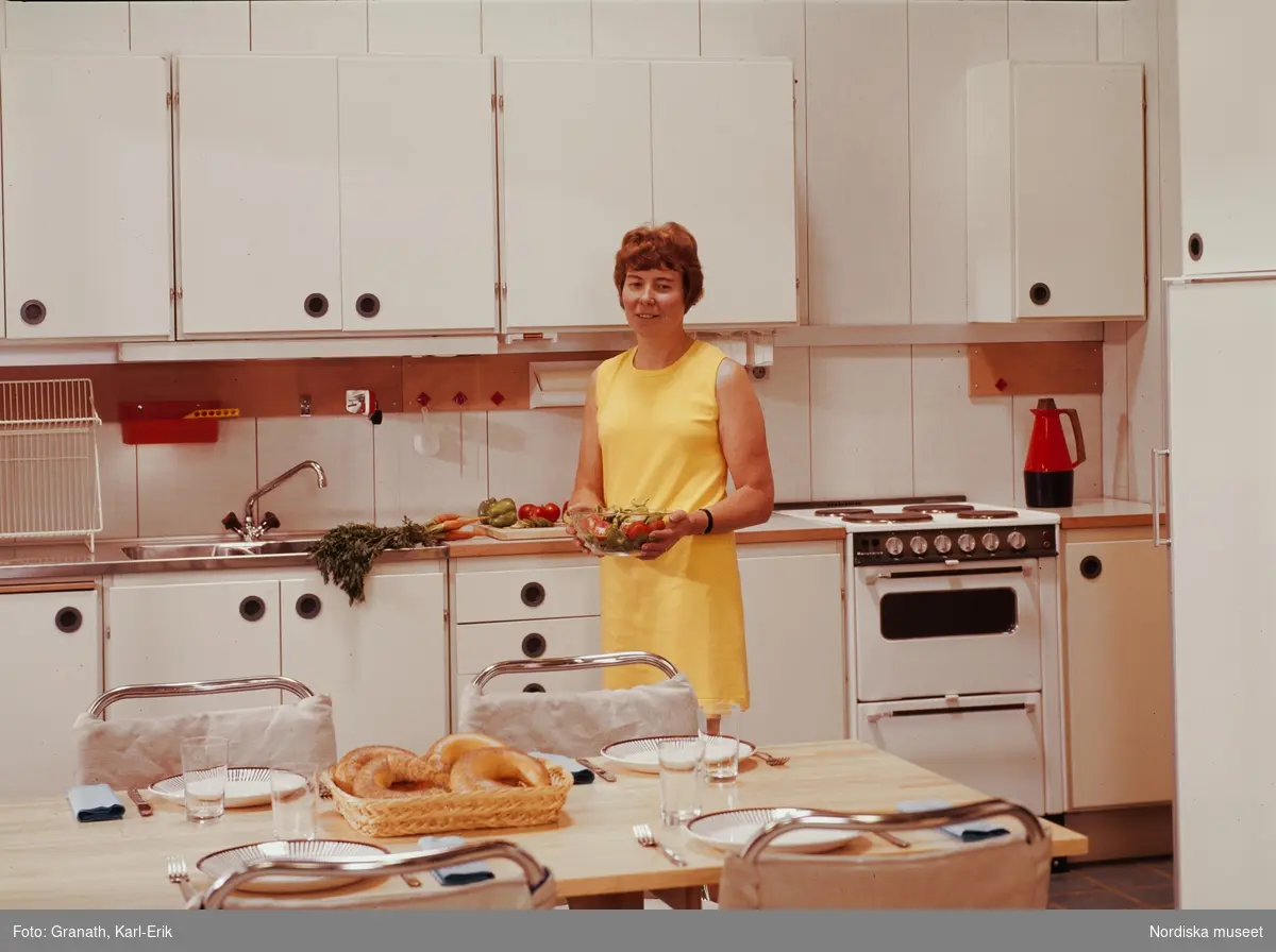 Köksinteriör. Kvinna i gul klänning med en salladsskål i handen