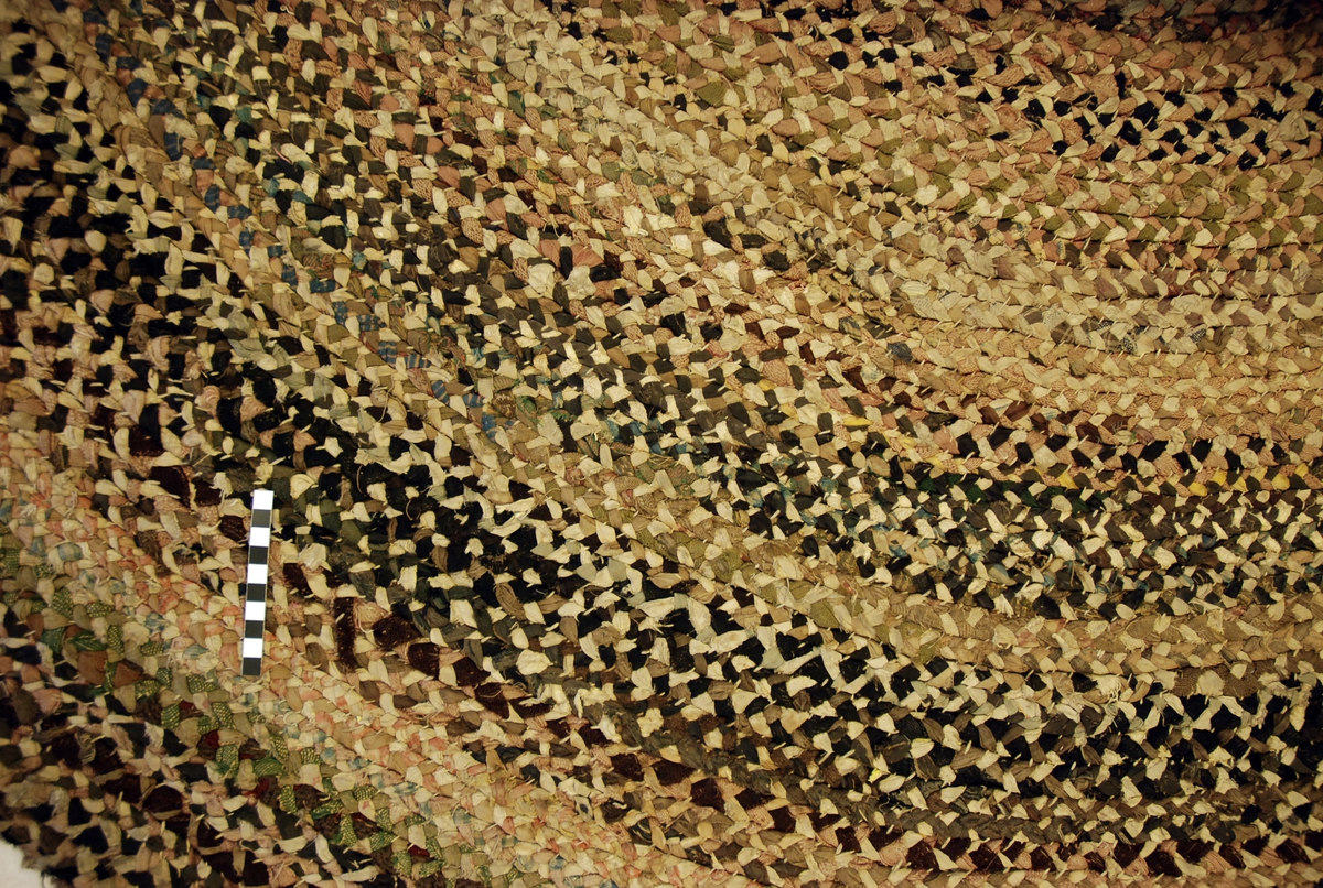 Rundt gulvteppe sammensydd av flettet tekstil. Flettingen består av 3 ulike tekstilremser som er skjøtet/sammensydd i endene. Fletteremsene er sydd sammen i en spiral med hvit bomullstråd på baksiden.