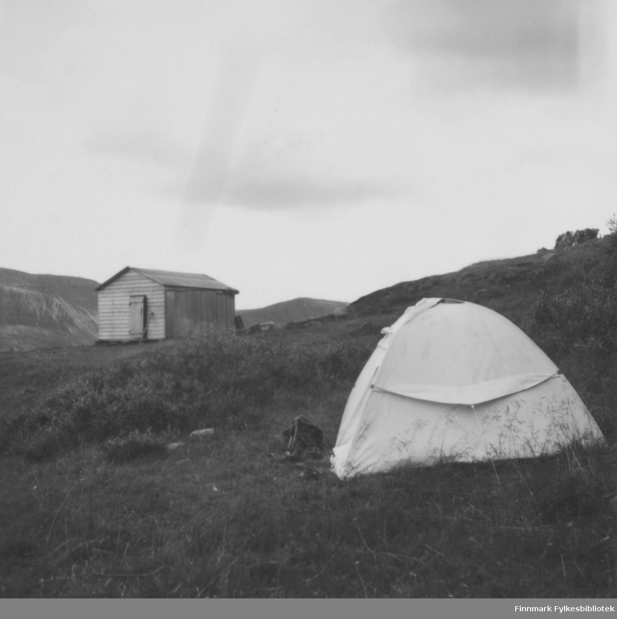 Ebeltofts telt slått opp på ukjent sted, kanskje i Finnmark. I bakgrunnen ei hytte, utenfor teltet en ryggsekk