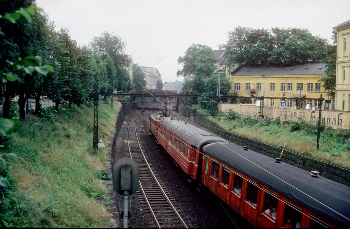 Persontog på vei inn mot Oslo V ved Strupen, området mellom Filipstad og Vestbanen. NSB elektrisk lokomotiv type El 11, vogner type BF1 og B22. Bygårder og plankegjerde med reklame for Sønnak batterier.