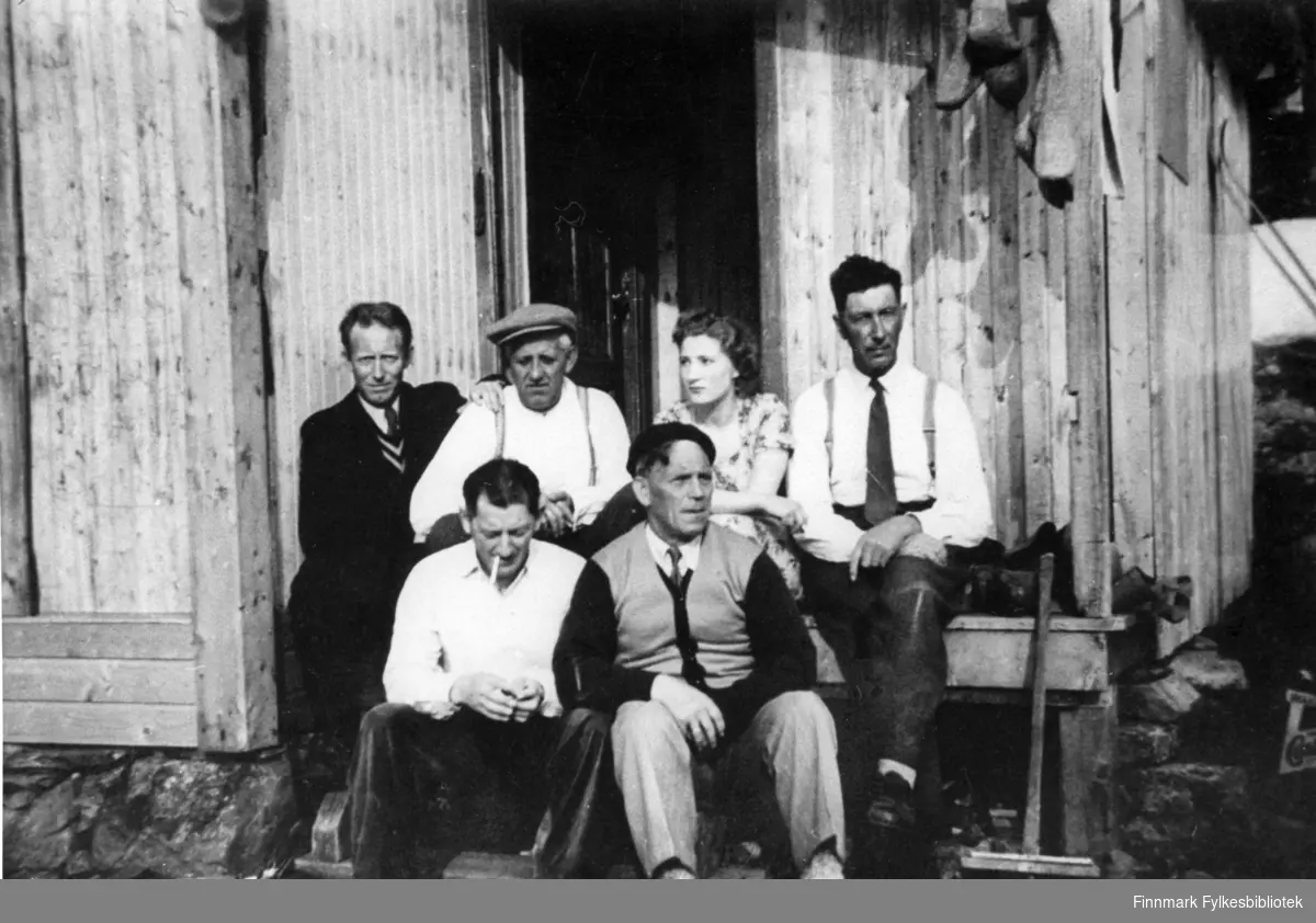 Portrettfotografi av en gruppe som sitter på trappa til en brakke. Forran fra venstre: Odin Nilsen og Håkon Iversen. Bak fra venstre: Agnar Iversen, Håkon Rapp, Sigrid Andersen og Odin Emaus.