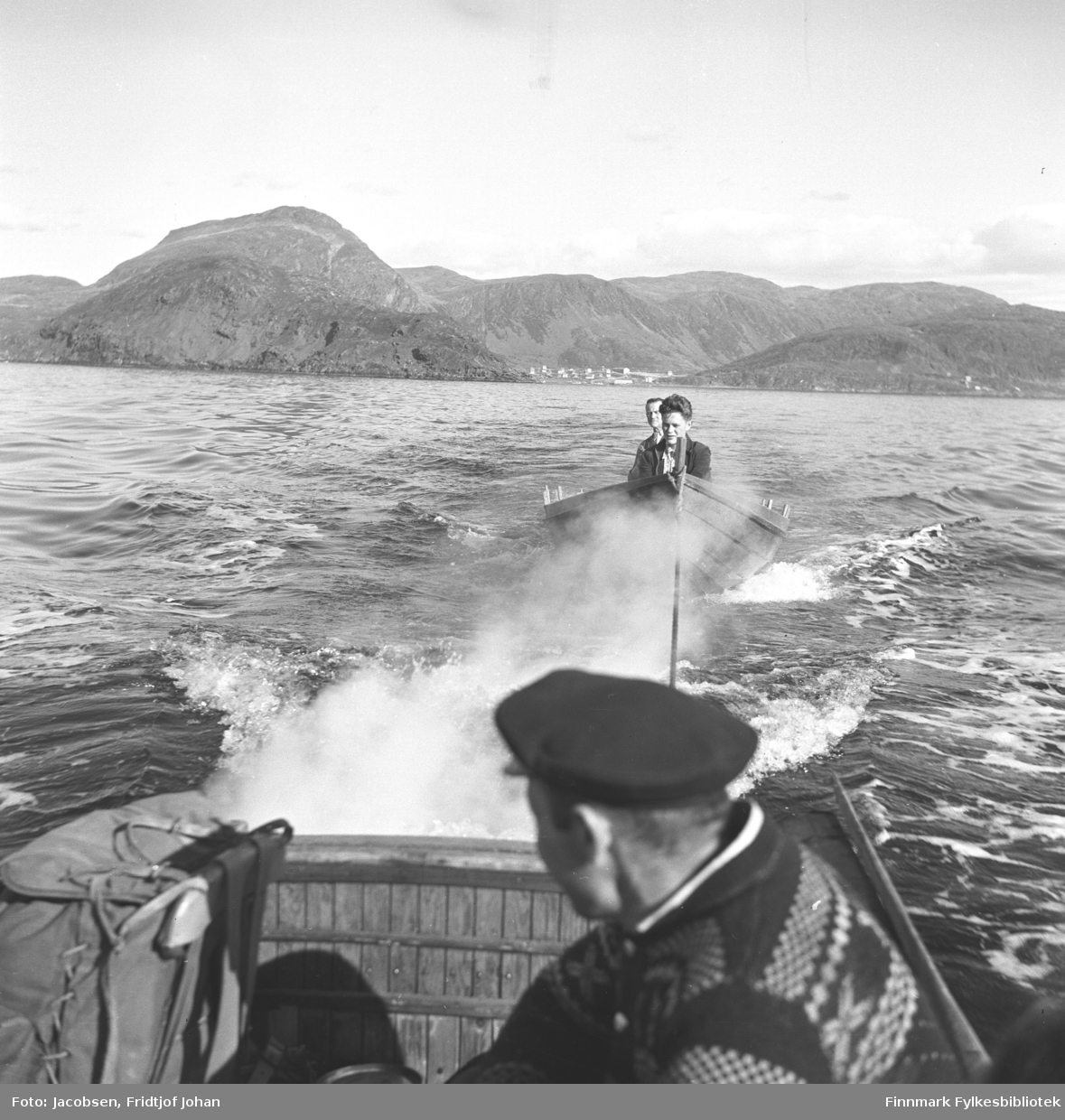 En robåt med to menn slepes av en liten motorbåt i Sørøysundet utenfor Hammerfest og Rypefjord. I motorbåten sitter en mann med strikkegenser, mørk lue og kikker bakover. I bakgrunnen mellom fjellene ligger Rypefjord. Til venstre på bildet ligger Rypklubben med Tyven i bakgrunnen.