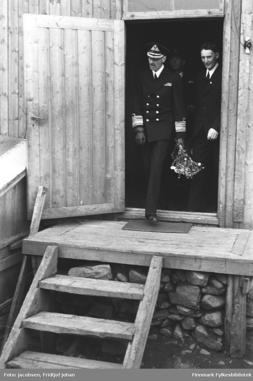 Kong Haakon VII på vei ut av lensmannsbrakka. Bildet er tatt under kongebesøket 10 juli. Han har uniform på seg og en blomsterbukett i sin venstrehånd. Litt bak han står en mann mørk dress og litt i bakgrunnen ses en mann i mørk uniform. Brakka er satt opp på en mur av stein og en liten matte ligger foran døra. Døra er kilt fast med en planke for å holde den åpen.