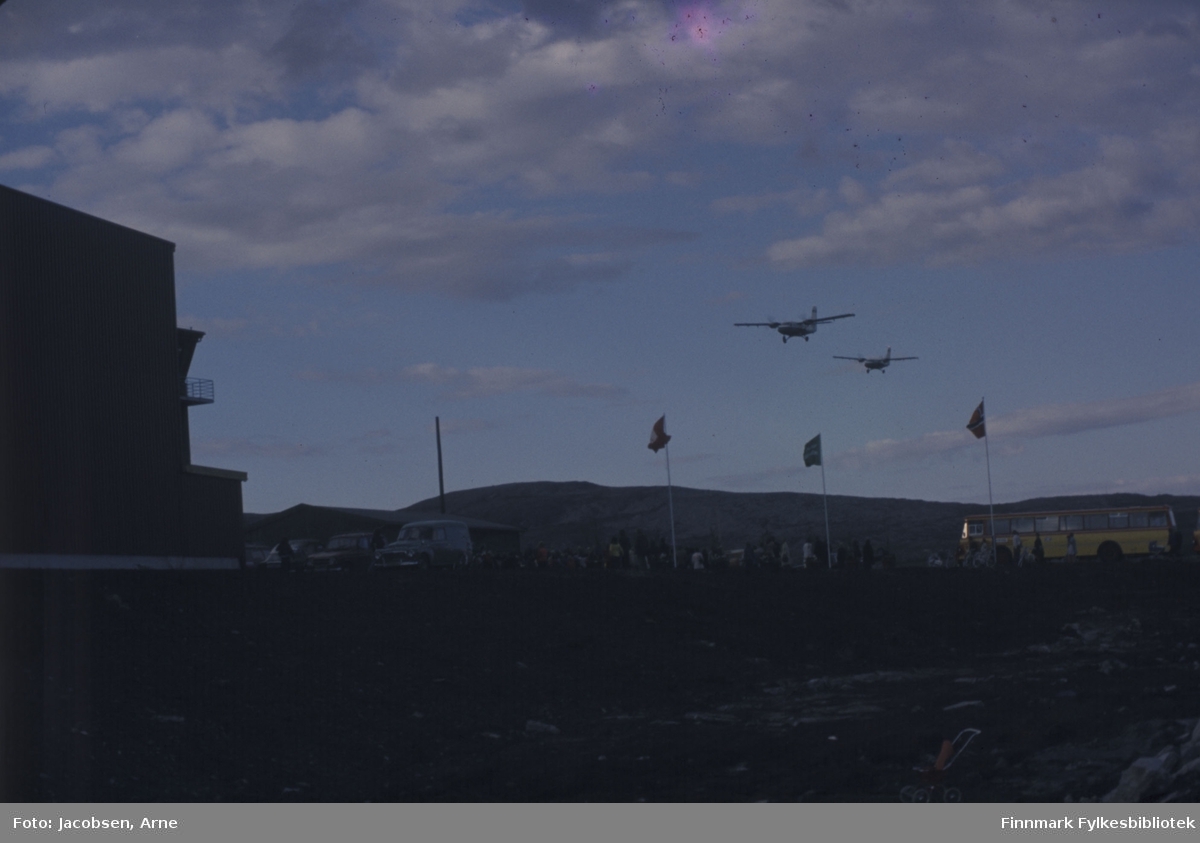 To Twin Otter-fly lander på Hammerfest lufthavn på åpningsdagen 30.07.74. Til venstre på bildet er hangaren og tårnet. Midt på bildet vaier Hammerfest- Widerøe- og det norske flagget. Til høyre står en FFR-buss som har fraktet noen av de mange menneskene som har møtt fram til markeringen. Terminalbygget ses bak de parkerte bilene. Delvis skyet og blå himmel over Reindalen som ses i bakgrunnen