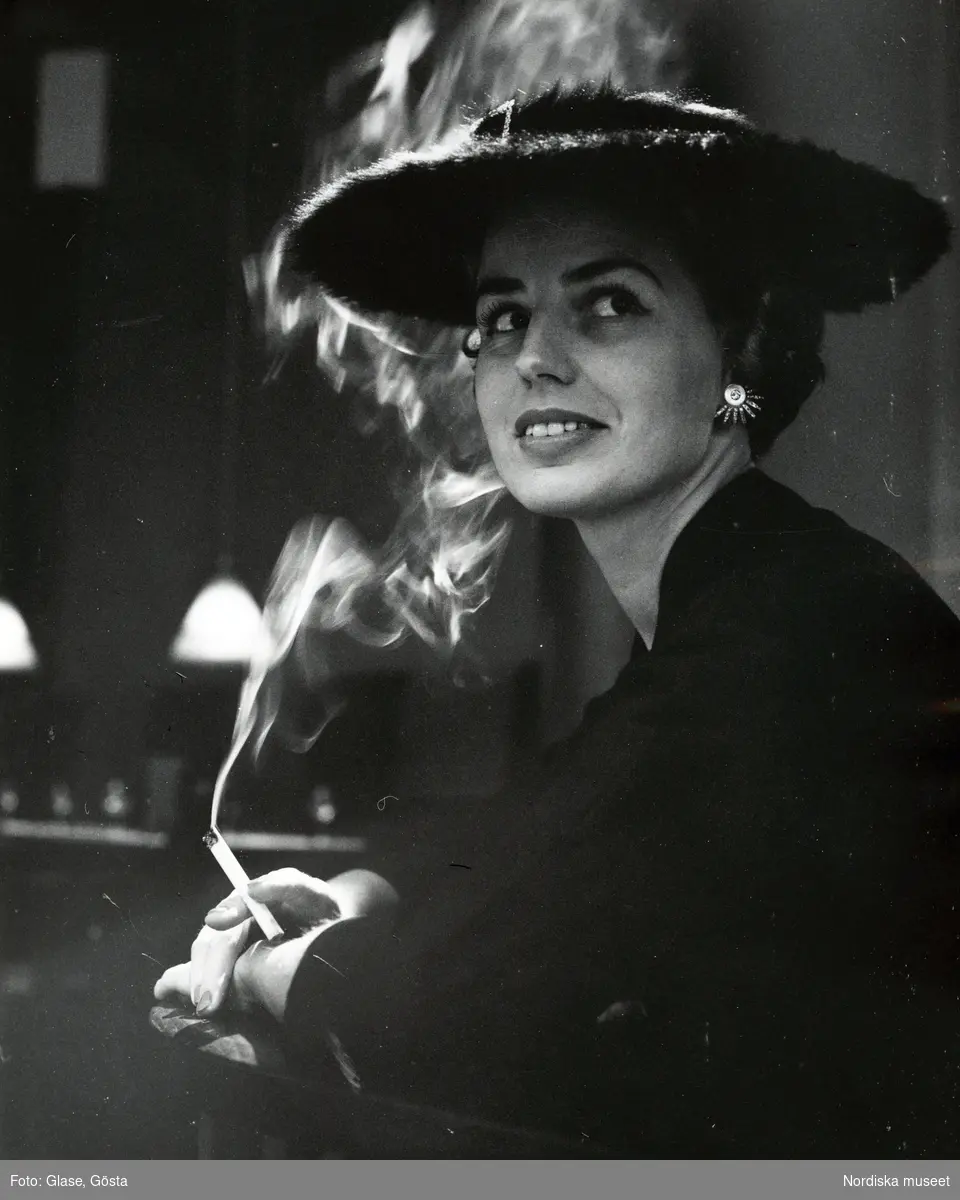 Porträtt av kvinna i hatt och örhänge med cigarett i handen.