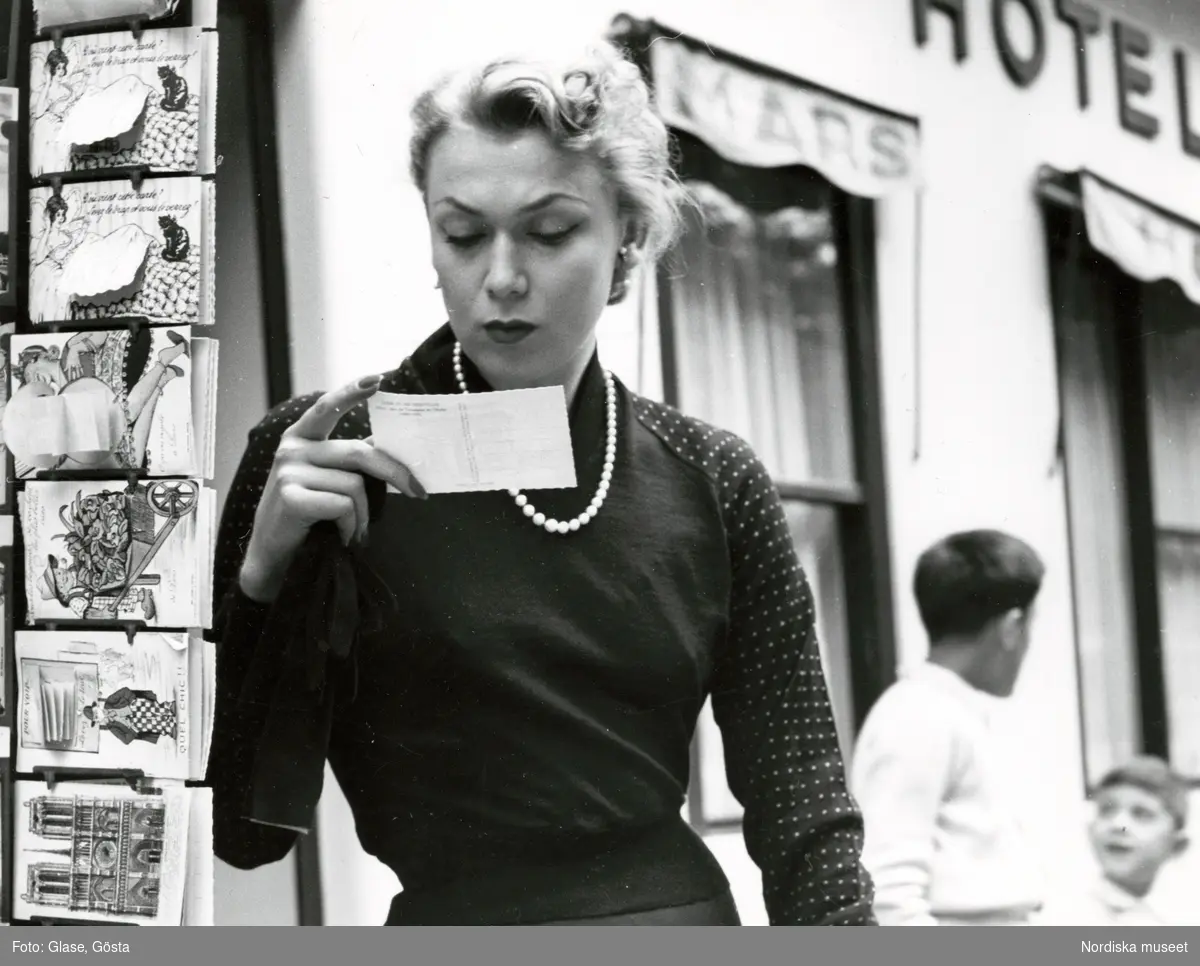Kvinna, mannekängen Anita Sandin, i tröja med prickar på ärmarna och pärlhalsband tittar på vykort.