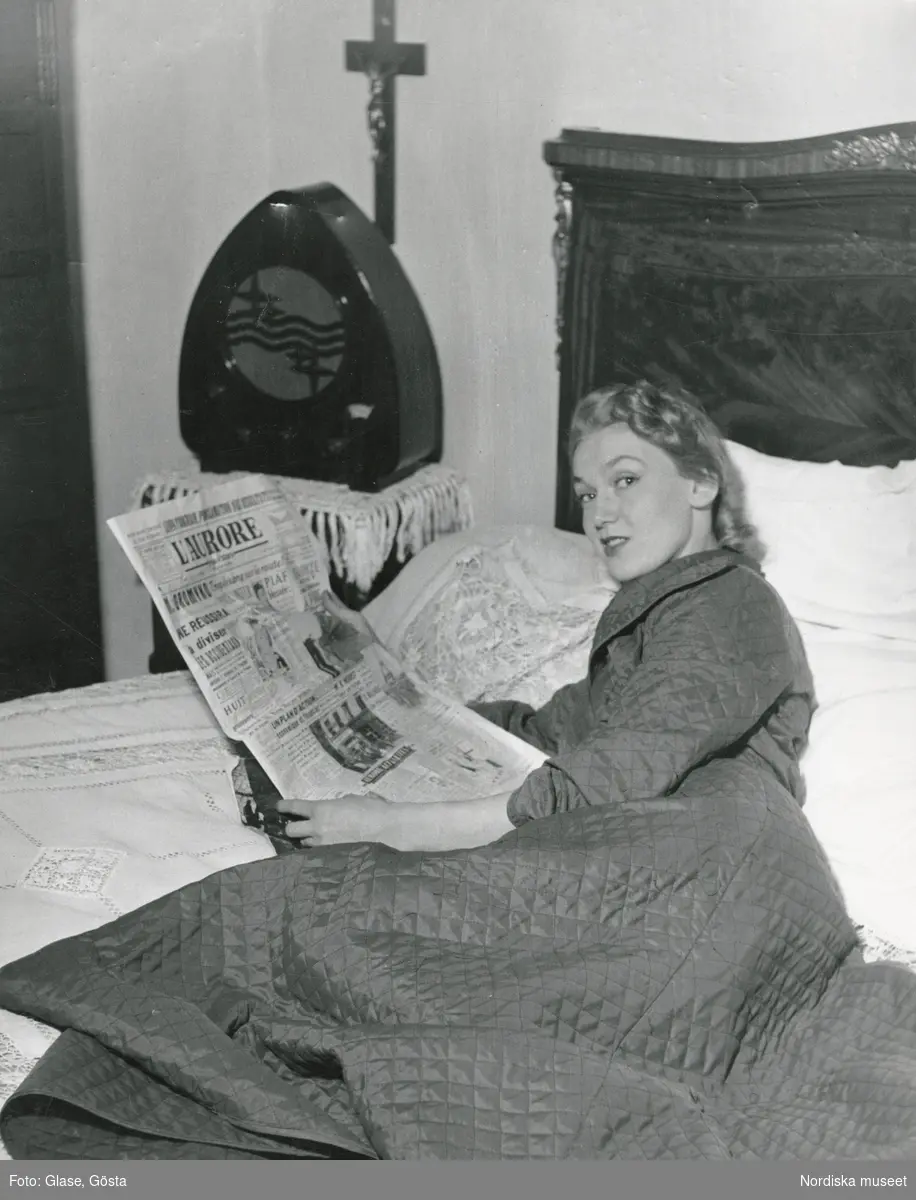 Kvinna, troligen mannekängen Anita Sandin, i quiltad morgonrock ligger på säng och läser tidningen L'aurore.
