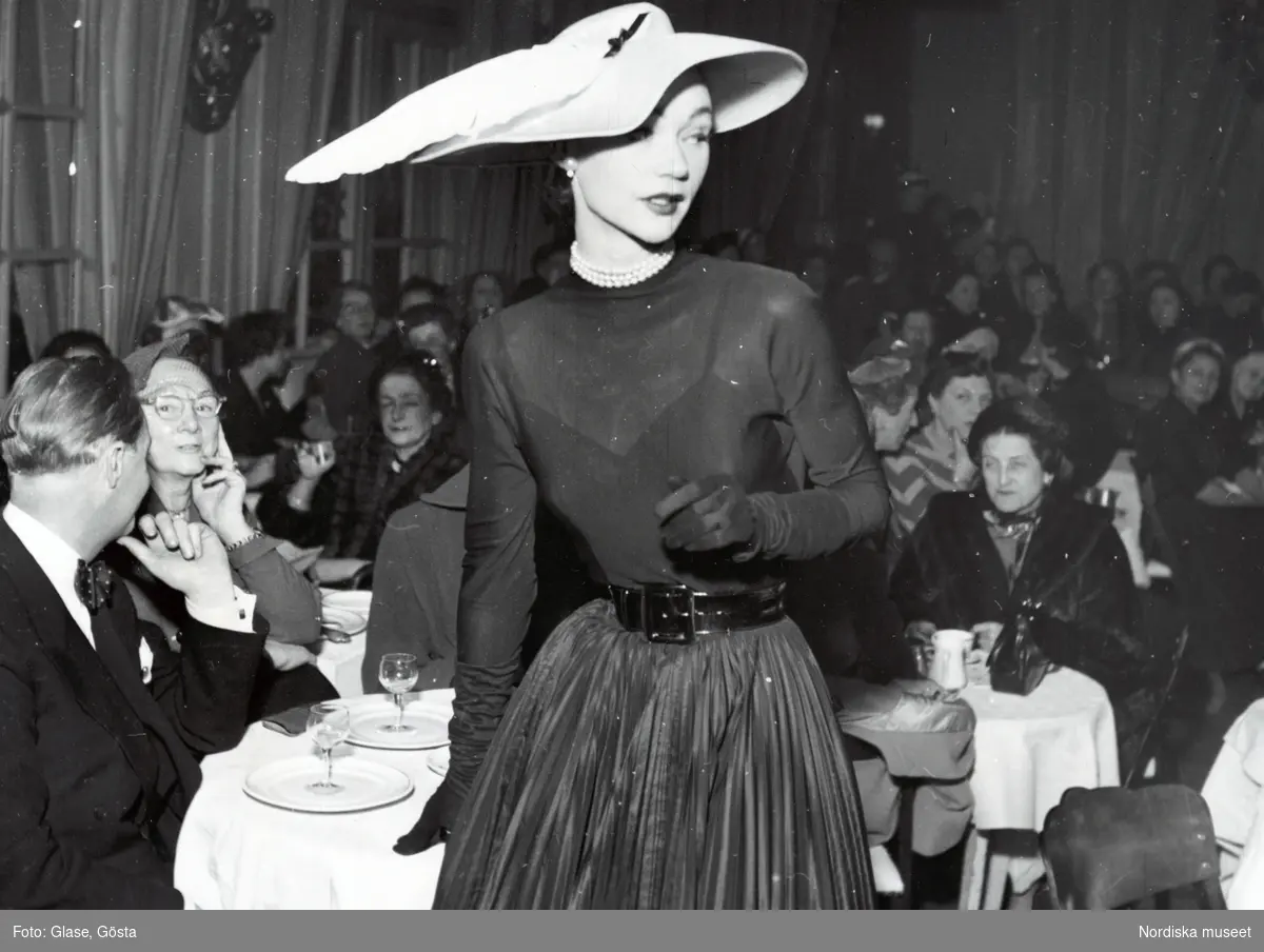 Modevisning - Modell i transparent tröja, handskar, plisserad kjol, lackskärp och hatt med fjäderpenna.